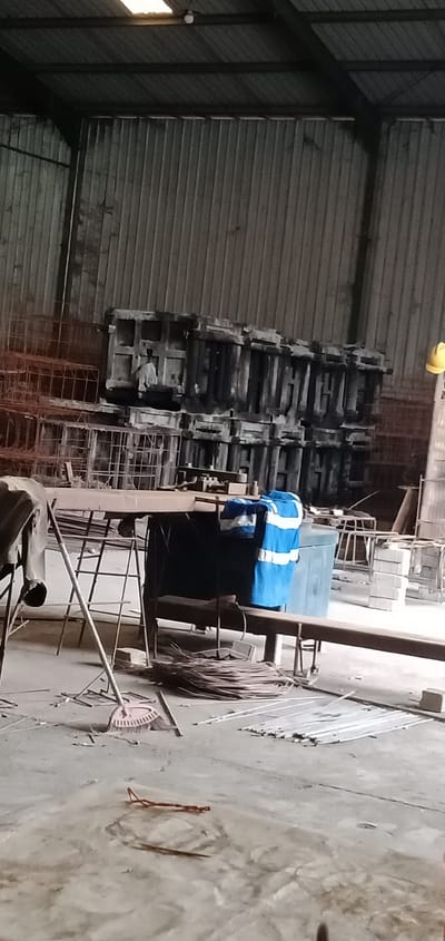 The image shows the interior of a warehouse or factory in Sumput, Indonesia. Corrugated metal walls and roof are visible. In the background, there are stacked concrete molds and rebar cages. A table is in the foreground with a blue and white garment on it. A broom leans against the table, and coils of wire are scattered on the floor. Scraps of metal and debris litter the concrete floor. A yellow hard hat is visible in the background.