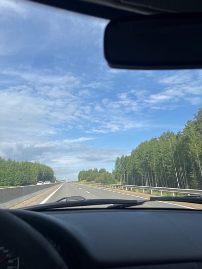 An image captured from inside a vehicle presents a view of a multi-lane highway stretching into the horizon, bordered by dense forests under a bright blue sky. The photograph is oriented sideways, but when viewed upright, it shows the interior of the car partially framing the exterior scene.

In the foreground, elements of the car's interior are visible. On the upper left, a portion of the rearview mirror is seen. On the lower right, the curve of a steering wheel and a section of the dashboard are present. The base of the windshield, including part of a wiper blade, is visible along the bottom edge of the frame.

The highway occupies the midground, featuring at least two lanes marked by white lines, indicating a clear path forward. A median strip or shoulder separates the road surface from the surrounding environment. A metal guardrail runs parallel to the highway on both sides. Tall, verdant trees form dense forests along the highway's edges, with some trees, particularly on the left, displaying light-colored trunks characteristic of birch trees.

The sky above is a vivid blue, populated by scattered, delicate white clouds, suggesting clear and pleasant weather conditions. In the distance, along the right side of the highway, several white vehicles, possibly vans or cars, are visible, either parked or moving slowly. Other vehicles can be faintly seen further down the road.

The scene depicts a daytime journey through a forested landscape. The location context indicates this is near Beloborodovo, Russia. The overall impression is one of open road travel amidst natural scenery during good weather. No discernible text is visible in the image.