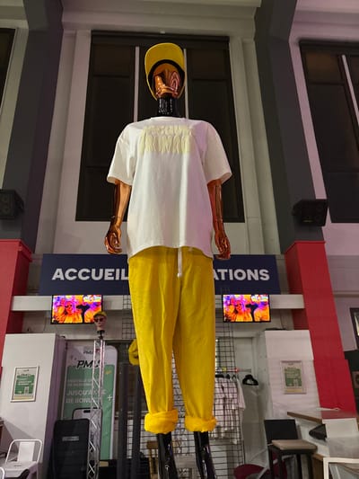 A mannequin sporting a yellow baseball cap, a white short-sleeved shirt with decorative ribbing, and yellow pants. The mannequin has a copper-colored complexion and black legs. In the background, screens display vibrant, colorful images. Signage with the word "ACCUEIL" is visible alongside an advertisement. A cluttered environment is apparent, including a metal rack and various displayed items.