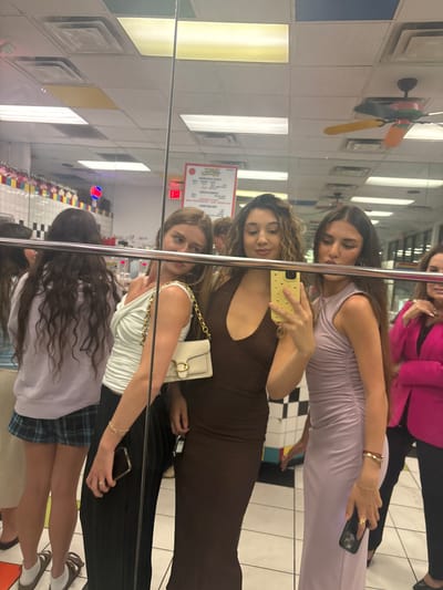 A mirror selfie captures five young women in a brightly lit indoor space, likely a restaurant in Boca Raton, United States, given the interior design elements. The woman in the foreground wears a light-colored top, a blue plaid skirt, and white socks with ballet flats. The second woman wears a brown dress and a white purse with a gold chain. The third woman wears a light purple dress. In the background, another woman wears a pink blazer. The environment features white tiled walls with colorful square accents, a menu board, and a whimsical decorative fan, typical of an In-N-Out Burger restaurant. The black and white checkered floor further indicates a classic diner aesthetic. The women are holding phones, suggesting they are taking a photo of themselves.