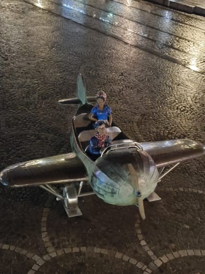 A metallic sculpture of a single-engine, high-wing monoplane is centrally positioned on a dark, wet cobblestone pavement. The aircraft features a visible front propeller and exhibits a greenish-blue patina on its forward fuselage and nose, with more reflective, possibly polished, wings and tail surfaces. Two stylized figures occupy open cockpits: a male-presenting figure in the front wears a blue cap and a red-patterned scarf, while a female-presenting figure in the rear wears a blue top and a red-patterned headscarf. The cobblestone ground reflects overhead artificial light sources, creating bright streaks, and incorporates patterns formed by lighter-colored stones. This sculpture is located at Place Saint-Lambert in Liège, Belgium.