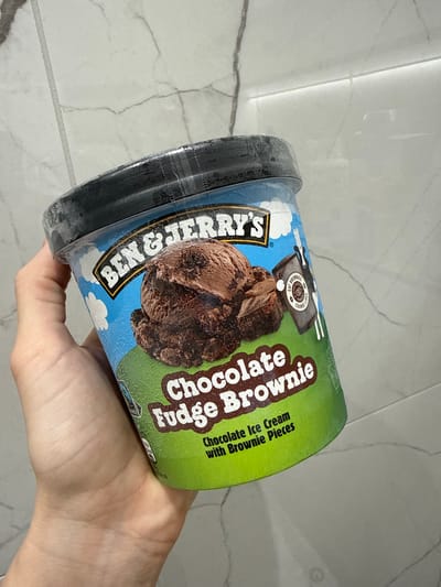 A hand holds a pint of Ben & Jerry's Chocolate Fudge Brownie ice cream. The container is angled slightly, showing the lid, the blue sky and cloud design, and the green portion with the flavor name. The background is a blurry, light-colored tiled surface.