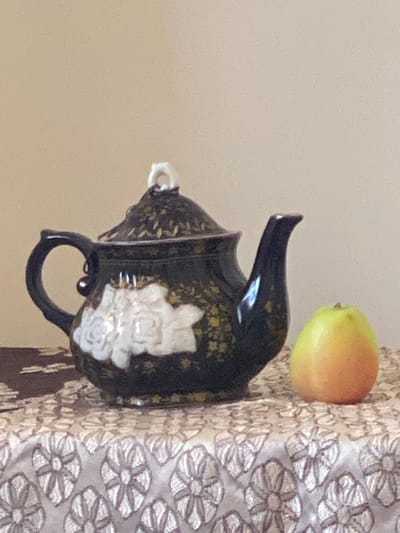 A still life arrangement is depicted on a textured tablecloth in what appears to be an indoor setting, potentially a home or a studio. The background is a plain, light-colored wall, devoid of any discernible features.
The primary subjects are a decorative teapot and a single piece of fruit. The teapot, dark in color and adorned with intricate gold patterns and white floral motifs, sits to the left of the frame. It has a curved handle, a spout, and a lid topped with a white finial. To the right of the teapot, a small, round piece of fruit, possibly an apple or a quince, displays a gradient of yellow and orange hues.
The tablecloth itself is a prominent element, featuring a repeating pattern of stylized, light-colored floral or starburst designs. To the left of the teapot, a darker fabric with a subtle pattern is partially visible.
The lighting appears soft and diffused, casting gentle shadows. There are no people present in the scene, nor are there any indications of specific activities or interactions taking place. The composition is static and focuses on the arrangement of the objects.
The image does not contain any contextual clues to suggest its location within Arbagish, Uzbekistan.