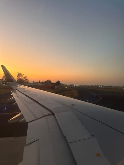The image displays a forensic view from a commercial aircraft window at an airport in Zapote del Valle, Mexico, during either sunrise or sunset. The lower left and central portions of the frame are occupied by the white wing of a Volaris aircraft, identified by the "volaris.com" branding on its winglet. The wing's flaps and control surfaces appear to be in a partially extended configuration, indicating the aircraft is on the ground, likely taxiing, preparing for departure, or having just arrived. To the right of the wing, a dark asphalt runway or taxiway stretches into the distance, marked by numerous illuminated blue edge lights and several yellow runway threshold lights further down the strip. The sky exhibits a prominent gradient, transitioning from a bright orange-yellow near the horizon to a lighter blue in the upper portion of the frame. In the background, beyond the airport perimeter, silhouetted trees, some indistinct buildings, and a notable red structure are visible against the illuminated sky. No individuals are discernible within the frame.