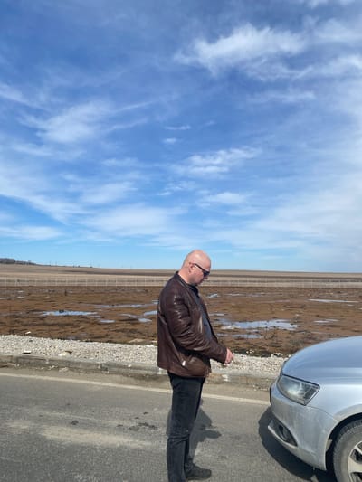 A bald man wearing sunglasses and a brown leather jacket stands on the side of a paved road, next to a silver car. He is looking out over a field with puddles of water. In the distance, a fence line runs across the field. Above, a blue sky is partially covered with white clouds. The location is Kliuchiki, Russia.