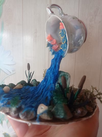 The image shows a miniature diorama depicting a waterfall flowing from an overturned teacup into a small pond. The teacup, which is white with floral accents, is positioned at an angle, with a blue, textured material representing the cascading water pouring out. Inside the teacup, there are small, coral-colored and blue decorative elements that resemble flowers or aquatic life.
The diorama rests on a silver-colored base, held by a person's hand, which is partially visible at the bottom of the frame. The scene includes various natural elements like rocks, pebbles, and green plant-like structures, some of which appear to be artificial reeds or grasses. There are also small, shell-like objects scattered around the pond area.
The background is a pale, textured wall, possibly wallpaper, with a subtle wood grain pattern. A portion of a large, light blue, stylized flower can be seen on the left side of the background, suggesting an artistic or decorative context. The overall impression is that of a crafted decorative item, possibly a tabletop display or a craft project. The lighting appears to be natural or from an overhead source, casting soft shadows.