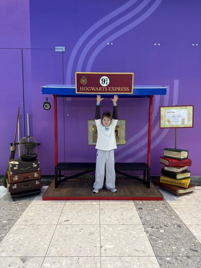A vertically oriented photograph shows a child posing at a "Hogwarts Express Platform 9 3/4" photo opportunity setup indoors. The child, likely a young boy, is wearing a white t-shirt and grey sweatpants, with his arms outstretched to grasp the top of the blue and red "platform." Behind the boy, there is a purple wall with light purple curved designs.
The setup includes a brown sign that reads "HOGWARTS EXPRESS" with the number "9 3/4" displayed above. To the right of the child are faux stacks of books, and above the books is a framed certificate. To the left of the child, there is a hanging "9 3/4" sign, stacked vintage suitcases, a birdcage, and a broomstick. The setup rests on a wooden platform with black supports.
The floor is covered with a tiled pattern of white and grey. The picture seems to be taken indoors, at a fan event location in Родниковское сельское поселение, Ukraine.