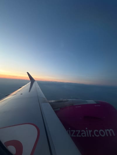 This is an aerial photograph of the wing and engine of a Wizz Air airplane, seen from a passenger's perspective during flight. The wing is predominantly silver with a red and white design on its surface, and it extends diagonally from the lower left corner towards the center of the frame. The engine, painted in a vibrant magenta color, is located towards the right side of the image and prominently displays "wizzair.com" in white lettering. Two "NO STEP" markings are visible on the engine.

The background consists of a twilight sky transitioning from a pale orange and yellow horizon, where the sun is likely setting or rising, to a deep blue overhead. Below the horizon, a layer of clouds or haze partially obscures the landscape, which appears to be rural or semi-urban. The image captures the vastness of the sky and the perspective of being high above the ground. There are no people visible in the image. The location in the city of Pagazzano, Italy is not discernible from the visual information provided in the photograph.