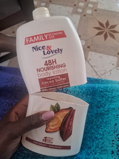In Muthambi ward, Kenya, a dark-skinned left hand with light pink manicured fingernails holds a white plastic "Nice & Lovely 48H NOURISHING body lotion" bottle. The outer layer of the bottle's label, which displays "FAMILY ECONOMY SIZE," "Nice & Lovely," "48H NOURISHING body lotion," "repairing cocoa butter and shea butter," and "visibly reduces stretch marks," is partially peeled back. This action reveals an inner label or surface on the bottle's lower section, which features "KENYA'S #1 BODY LOTION," "NON GREASY - NON STICKY" text, and an illustration of cocoa pods with leaves. The hand and bottle are positioned over a blurred blue and purple patterned fabric. In the background, a light-colored tiled floor with a repeating star pattern is visible, along with a dark, indistinct piece of furniture in the upper left.