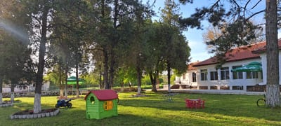 The image shows a grassy lawn area with several trees, a building, and children's play equipment. The building is white with a red tile roof and several windows. There is a green umbrella outside the building. Several trees are scattered across the lawn, some with whitewashed trunks. Children's play equipment includes a green and red playhouse, a blue motorcycle, and a small red table with chairs. There is also a bicycle leaning against a tree trunk. The scene appears to be set in Zlatina, Bulgaria, as indicated by the language on the umbrella, which is consistent with Bulgarian.