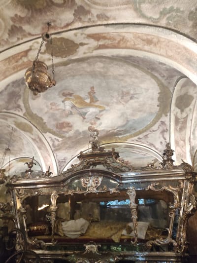 The image depicts the reliquary of Saint Julius inside the Basilica of San Giulio in Orta San Giulio, Italy. The reliquary is an ornate, silver-plated glass casket containing the mummified body of the saint, dressed in ornate vestments and lying on a velvet cushion. The casket is adorned with intricate carvings and decorative elements. Above the reliquary, the arched ceiling of the basilica is decorated with frescoes, featuring religious scenes with figures and cherubs. A gilded lamp hangs from the ceiling, casting a warm light on the scene. The overall atmosphere suggests a sacred and historical setting.