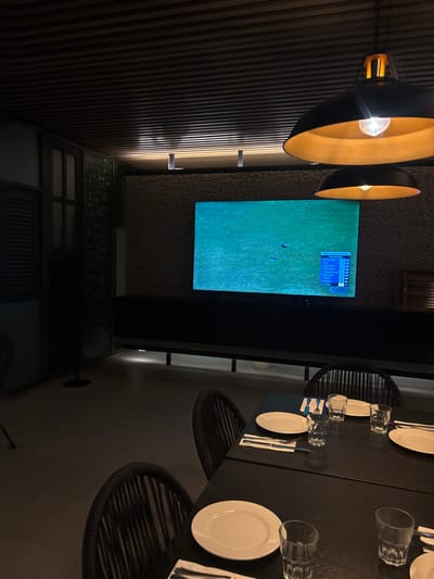 A modern dining area features a dark wood table set with white plates, glasses, and cutlery. Wicker-style chairs surround the table. A television screen displays a golf broadcast, positioned between the table and a wall with vertical wood panels. Two pendant lights with gold interiors hang above. A lower section of the wall includes a window or decorative panel.