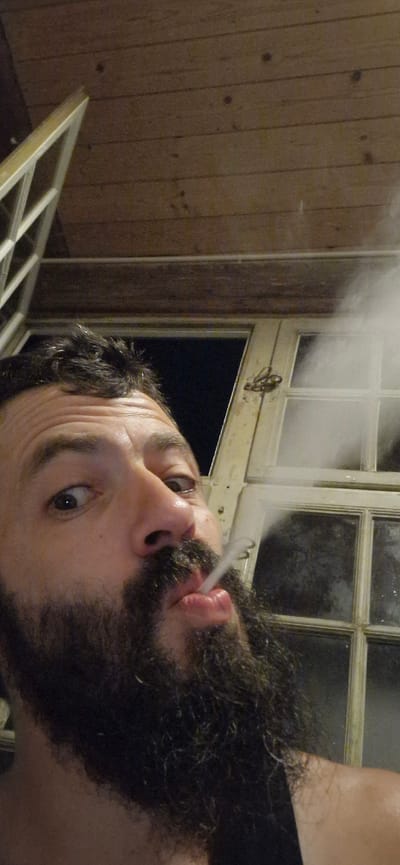 A man with dark hair and a full beard is exhaling smoke. He is looking towards the camera with a surprised expression. The background shows a wooden ceiling and a window with multiple panes. The overall lighting suggests it is either night or a dimly lit interior. The man appears to be indoors, possibly in a rustic setting given the wooden elements and the style of the window. The image seems to be a selfie. The scene is set in Wattenwil, Switzerland.