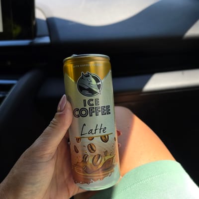 A hand with a light pink fingernail and a small cut on the thumb holds a gold and light blue can of "ICE COFFEE Latte" with coffee beans depicted on it. The can is positioned in the foreground, with the hand obscuring part of it. The background shows a dark interior, likely a car, with a bright highlight on a surface, possibly a dashboard or window. The scene appears to be outdoors during the day, as indicated by the sunlight. There is no identifiable location in the city of Ravnogor, Bulgaria.