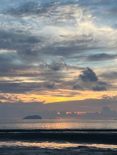 The image depicts a serene sunset over the ocean, likely taken from a beach in Cuyo, Philippines. The sky is a canvas of dramatic clouds, transitioning from pale blues and greys at the top to warm oranges and yellows closer to the horizon. The setting sun is partially obscured by dark clouds, casting a golden glow on the water's surface. In the distance, a small, uninhabited island stands silhouetted against the vibrant sky. The foreground shows a wet, sandy shore, reflecting the colors of the sunset. No people or specific structures are visible, emphasizing the natural beauty of the scene.