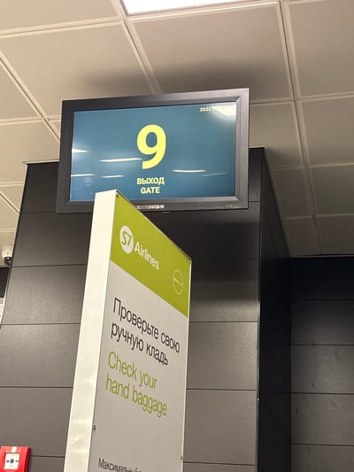 This image depicts an indoor airport or transit gate area in Bol'shie Kabany, Russia.
A prominent black-framed digital display screen shows a large yellow '9' centered on a blue-green background. Below the number, 'ВЫХОД' (Exit) and 'GATE' are displayed in white text. The upper right corner of the screen indicates '23:21 06'. The bottom edge partially shows 'RANSINFORM'.
To the lower right of the screen, a white and green S7 Airlines freestanding sign is angled towards the viewer. The sign features the S7 Airlines logo and the 'one world' alliance logo. Text in both Russian ('Проверьте свою ручную кладь') and English ('Check your hand baggage') instructs passengers to verify their hand luggage. A portion of additional Cyrillic text, 'Максимальный', is visible at the bottom.
The background consists of dark grey, horizontally paneled walls. A white dome-shaped security camera is mounted on the wall to the left. A red fire alarm pull station is partially visible at the bottom left. The ceiling is a grid of light grey, perforated acoustical tiles with integrated rectangular fluorescent light fixtures. No people or active interactions are present in the scene.