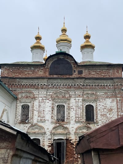 The image shows the exterior of an old, weathered, white-washed brick church with a red tiled roof and golden domes. The church features multiple arched windows, some with bars and broken glass, and decorative brickwork. The facade is partially covered by a dark, weathered metal sheet. In the foreground, there are two dark, sloped roof structures made of corrugated metal, partially obscuring the lower part of the church. The sky is overcast and gray. The scene appears to be in the village of Кстинино, Russia, indicated by the context provided. There are no people visible in the image.