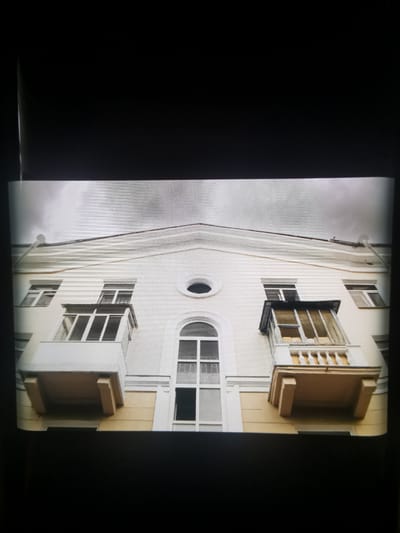 This media file depicts the facade of a building, likely an apartment block, viewed from a low angle looking upwards. The architecture suggests a style common in Eastern Europe, with a pale yellow and white color scheme. The building has multiple windows and two balconies are prominent. The balcony on the left is open with white railings and a light-colored platform. The balcony on the right is enclosed with glass panes and has a dark frame. A large, arched window with multiple panes is centrally located, and a smaller, circular window is visible above it. The sky appears overcast and gray, suggesting a cloudy or possibly rainy day. The building is situated in a residential area, as indicated by the apartment-style balconies and windows. There is no visible text in the image. The scene is static, with no people or ongoing activity, focusing solely on the architectural elements of the building. The lighting suggests it is daytime, but the cloudy weather creates a subdued atmosphere. The location is identified as Serov, Russia.