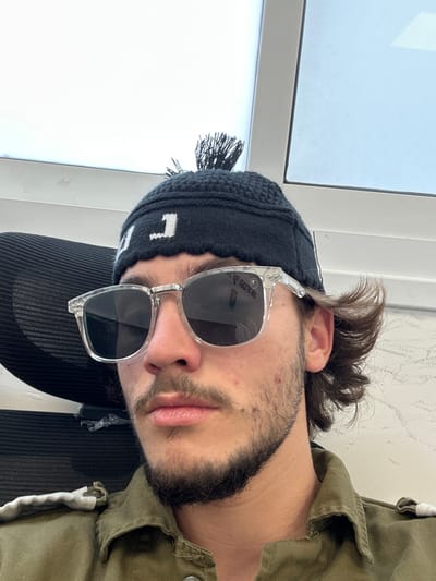 A close-up image features a male individual centrally framed, looking directly forward. The individual wears a black knitted beanie with a white "J" character and a small black pom-pom, and clear-framed sunglasses with dark lenses. Visible facial features include a mustache and beard stubble, and minor skin blemishes on the cheeks. Dark brown hair is visible on the sides of the head beneath the beanie. The individual is wearing an olive-green or khaki collared garment, possibly a shirt or jacket, with a visible snap button closure and a light-colored strap detail on the left shoulder. A black mesh chair is partially visible behind the individual's left side. The background is a bright, light-colored indoor space featuring white vertical and horizontal architectural elements, likely window frames, indicating natural light. The specific location within Galil Tachton Regional Council, Israel, is not discernible from the image.