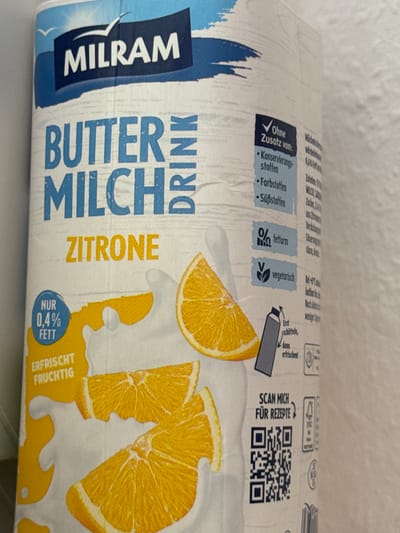 This is a close-up, slightly angled shot of a Milram "Buttermilch Zitrone" (buttermilk lemon) drink carton. The carton has a white background with blue and yellow graphics. The Milram logo is prominently displayed in the bottom left corner in blue with white lettering. Large blue text on the carton reads "BUTTERMILCH ZITRONE."

Several informational icons and text snippets are visible:
- A blue checkmark with the text "Ohne Zusatz von:" (Without added:) followed by bullet points listing "Konservierungsstoffen" (preservatives), "Farbstoffen" (colorants), and "Süßstoffen" (sweeteners).
- An icon indicating "fettarm" (low fat) with "%".
- A green "V" indicating "vegetarisch" (vegetarian).
- A graphic of a lemon slice with a yellow splash and text reading "NUR 0,4% FETT" (Only 0.4% fat) and "ERFRISCHT FRUCHTIG" (Refreshes fruity).
- Text suggesting to shake the carton ("Erst schütteln, dann erfrischen!").
- A QR code with the text "SCAN MICH FÜR REZEPTE" (Scan me for recipes).

The image appears to be taken indoors, likely in a kitchen or pantry setting, given the smooth, light-colored background which could be a wall or countertop. There are no people or animals in the image. The lighting is neutral, suggesting artificial light or an overcast day. The overall impression is one of a clean, modern product packaging.