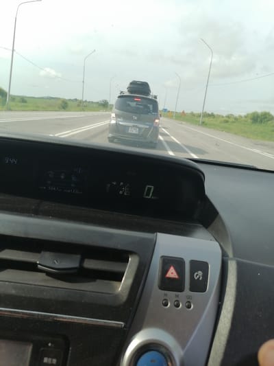 The image is a first-person view from inside a car driving on a highway. The dashboard displays "9:44" at the top left and the trip meter shows "504.1 km" with an average fuel consumption of "12.0 km/L". To the right, the gear selector is visible, showing "P R N D B". In the center display, a diagram of the car's hybrid system is shown. The car in front is a dark-colored minivan with a roof rack. The road is asphalt with white lane markings, and there is a grassy shoulder with trees and bushes. The sky is overcast. The location is identified as Aeroport, Russia, based on the license plate of the car in front, which reads "O 460 EE 78".