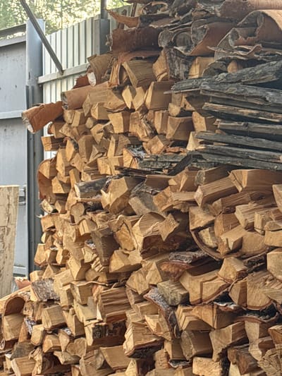 A large, unsheltered pile of cut firewood is stacked against a corrugated metal fence. The firewood consists of numerous split logs and some bark pieces, varying in size and shape, with a predominantly brown and tan color palette. The fence, a dull gray, appears to be part of an industrial or utility area. The background shows faint greenery and sunlight, suggesting an outdoor setting, possibly a yard or storage area within the city of Ust-Mana, Russia, though no specific landmarks confirm this location. The image captures a still, inanimate scene with no people or discernible actions.