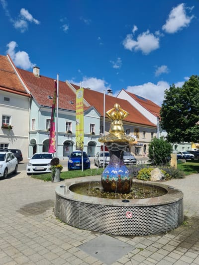 The image captures a public square in Sankt Marein bei Neumarkt, Austria, under a bright blue sky with scattered white clouds. A prominent fountain with a gold-colored sculpture, possibly depicting a seated figure, stands at the center of the scene. The base of the fountain is adorned with colorful mosaic tiles and has a circular, perforated metal surround. Water cascades from the sculpture into the fountain's basin.

Surrounding the fountain are paved walkways and a small landscaped area with green plants and rocks. In the background, a row of traditional Austrian buildings with red-tiled roofs lines the square. These buildings feature light-colored facades, windows, and architectural details typical of the region. Flags and banners are visible in front of some of the buildings, one of which appears to be a yellow banner with text and an illustration of a bee.

Several cars are parked along the street in front of the buildings, including white, blue, and silver vehicles. The overall atmosphere is that of a sunny day in a charming European town. The image does not show any people actively interacting with the fountain or the environment.