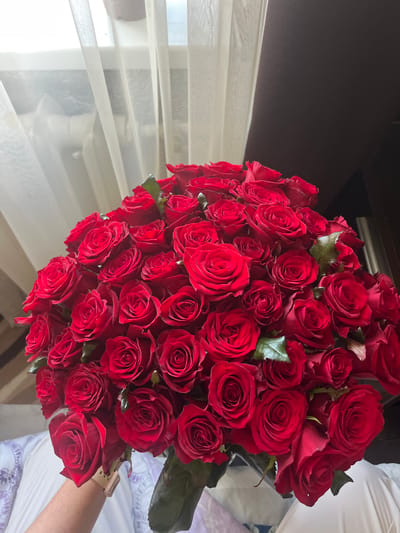 A bouquet of red roses is held by a person's arm, whose left hand is visible wearing a watch. The bouquet is large and fills the majority of the frame, with the roses being a deep red color and tightly packed together. Behind the roses, sheer white curtains are visible, obscuring a window. To the right of the curtains, a dark, textured surface is present, possibly furniture. The overall lighting suggests an indoor setting, likely a room. The image does not contain any specific landmarks or elements that would indicate a location within Vasylkiv, Ukraine.