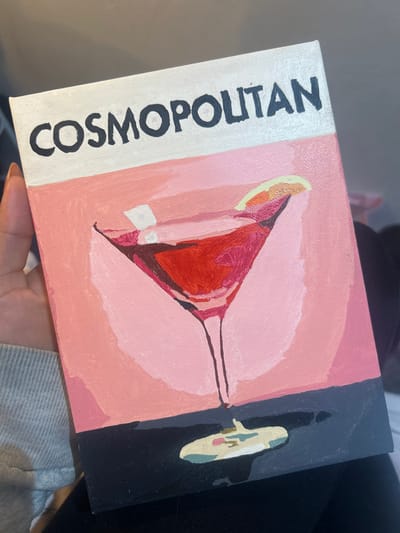 A rectangular canvas painting is held vertically by a person's left hand. The painting features a stylized depiction of a Cosmopolitan cocktail. The top portion of the canvas has a white horizontal band with the word "COSMOPOLITAN" written in black capital letters. Below this, the main background is light pink, transitioning to a slightly darker shade on the right. A martini glass, containing a red liquid and two small white elements, is centrally depicted. A wedge of citrus, yellow and orange, rests on the rim of the glass. The glass has a thin stem extending downwards to a dark grey or black horizontal band at the bottom of the canvas. On this dark band, beneath the glass, is an irregular, multicolored puddle, featuring light green, beige, and pink tones, implying a spill. Brushstrokes are visible across the painted surface, indicating an acrylic or oil medium. The person's hand, exhibiting light skin and short fingernails, grasps the left edge of the canvas. A light grey, textured sleeve covers the person's forearm. The background is dark and out of focus, with a hint of dark fabric in the lower right, providing no specific environmental details. The location of this scene in Winslow, United Kingdom, cannot be determined from the provided image content.