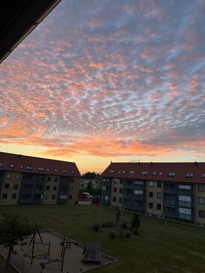 The image depicts a residential scene at dusk in Roskilde, Denmark. The sky is filled with vibrant, patchy clouds tinged with pink and orange hues, characteristic of a sunset. Two apartment buildings, featuring red tiled roofs and light-colored facades with dark-trimmed windows and balconies, dominate the middle ground. The buildings are set back from a grassy courtyard, which contains a children's playground with a sandpit, swings, and climbing structures. Several trees are visible within the courtyard and behind the buildings. The lighting suggests it is late evening, with shadows beginning to lengthen across the grass. No people are visible in the frame. The overall atmosphere is serene and domestic.