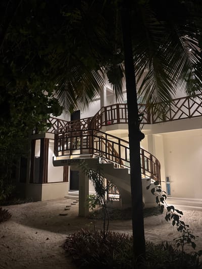 The image displays a multi-story building, identified as "AILA HOUSE 08," at night in Himmafushi, Maldives. The structure is predominantly white with a modern design, featuring an external, prominent spiral staircase constructed from white concrete steps with dark wooden railings and balusters. These railings exhibit both vertical slat and crisscross patterns. The staircase ascends to at least two upper levels, each with balconies or walkways also featuring dark wooden railings. A room number "401" is visible on an upper floor.
The ground surrounding the building is composed of light-colored sand. Flat, light-colored stepping stones are embedded in the sand, forming a pathway towards a ground-floor entrance. The environment is tropical, characterized by dense vegetation, including a large palm tree trunk in the foreground with fronds extending into the upper frame. Various smaller shrubs and plants, some with reddish foliage, are positioned around the base of the building and along the sandy ground.
The scene is illuminated by artificial light sources emanating from within the building and external fixtures, casting a warm glow on the white walls and staircase. No individuals or active subjects are present in the image.