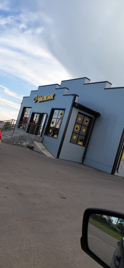 The image shows a blue, corrugated metal building with a stepped facade, decorated with yellow lettering spelling "ЧИЖИК" and window decals resembling cartoon eyes. The building appears to be leaning at an angle, creating an optical illusion. The entrance features a set of stairs and a ramp. In front of the building, there is a paved area, a trash receptacle with flowers, and a red car partially visible on the left. The sky is partly cloudy with visible cloud formations. The reflection in the car's side mirror shows a road with trees and greenery. This building is located in the city of Agapovka, Russia.