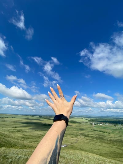 In Andreevka, Russia, a person's arm and hand extend upwards, palm facing the viewer, against a backdrop of a vast, blue sky dotted with white, fluffy clouds. The hand is adorned with white, almond-shaped fingernails and a silver, wave-like ring. A black smartwatch is visible on the wrist. Below the sky, a verdant, rolling landscape stretches out, featuring green fields, distant trees, and a small village with buildings. A winding dirt path is visible on the hillside, and a few cars are parked near it. The lighting suggests it is daytime, likely late morning or early afternoon, with shadows casting across the landscape.