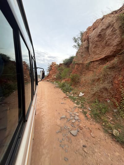 The image captures a view from inside a moving vehicle, likely a bus, traveling along a dirt road beside a rocky, reddish-brown cliff face. The perspective is from the side window, looking outwards and slightly upwards. The cliff is covered in sparse vegetation, and there is some litter visible on the ground near the cliff. The road ahead appears to be a track with loose stones and sand.
The time of day seems to be overcast or perhaps early morning, with a bright, diffused sky. The weather appears to be dry. The landscape suggests a rural or semi-arid environment. The bus itself is visible in the foreground, with its windows reflecting the surrounding scenery. A side mirror of the vehicle is also visible, showing a reflection of the road and the cliff. The image is shot with a wide-angle lens, creating a slightly distorted perspective. There is no visible text in the image. The overall impression is one of travel through a rugged and natural landscape.