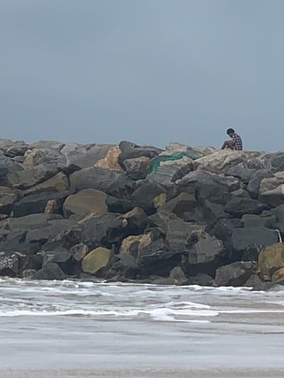 This image captures a coastal scene in Ibeju, Nigeria, under an overcast sky. The foreground shows agitated water with white foam, indicating ocean waves. Dominating the mid-ground is a massive, irregularly constructed barrier composed of numerous large, angular boulders in varying shades of grey, brown, and tan. A single human figure is seated atop the upper section of this rock barrier, positioned towards the right side of the frame. The individual wears a checkered shirt and dark lower garments, facing away from the camera, appearing to observe the water or horizon. To the immediate left of the seated person, a segment of green netting or fabric is draped over the rocks. No other individuals or significant structures are visible.