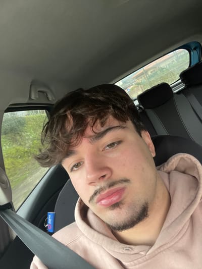An image taken from inside a vehicle depicts a young adult male in the foreground. He has dark, wavy hair, light brown eyes, a mustache, and a goatee. He is wearing a light peach-colored hooded sweatshirt and is looking directly at the camera. A black seatbelt is visible across his chest, buckled. To the lower right of his face, a small blue cylindrical object with "SPARCO CORSE" branding and a black lanyard is visible near the seatbelt connection point. The vehicle's interior features a light grey headliner and black rear seat headrests. Through the vehicle's windows, a blurred exterior environment is visible, indicating motion. Green vegetation is prominent on the left, while the rear window shows houses with light-colored walls and tiled roofs, interspersed with more greenery and a visible utility pole or street sign. The specific location within Santa Marta de Penaguião, Portugal, is not discernible from the visible external features, but the environment suggests a residential or semi-urban road.