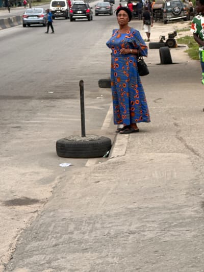 A woman in a blue and orange patterned dress stands on a street with cars and people in the background. A black purse hangs from her shoulder. In the foreground, a tire sits on a metal pole, seemingly embedded in the road. The road surface is grey and shows signs of wear. More tires are visible further down the road.