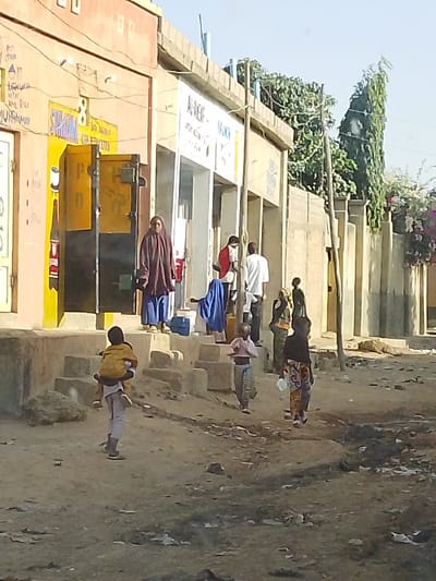 The image captures an outdoor scene, likely a street or public area in a town or village under bright daylight. On the left, there are two connected buildings. The first building, closest to the viewer, is painted a light orange or peach color with a prominent yellow section around its two open doorways. The interiors of these doorways appear dark. Above the left doorway, faint text is visible, including "STAR," "LEGADA MTN," "MA," "KAI RICE," and "MUHAMMAD." Vertically along the left door frame, "DPO" can be read. The second building to its right is lighter, possibly white or light gray, and also has open entrances. Text on this building's facade reads "AL-AKAF - ADANCA." Both buildings have flat roofs with a low parapet wall, and some utility wires are visible against the sky near the first building.
A set of concrete steps leads up to the entrances of these buildings from the dirt ground below. Several individuals are present in the scene. An adult woman, wearing a long, dark purple or maroon garment and a head covering, stands on the steps in front of the first building, looking generally towards the left. Near the second building's entrance, a man in a red shirt and white trousers is partially visible, along with another man in a white shirt and dark trousers. A blue piece of fabric is draped over something on the ground near them.
In the foreground, on the unpaved, dusty ground, several children are visible. To the left, one child is carrying a smaller child on their back in a piggyback ride. Both are wearing light-colored clothes and walking towards the right. Further to the right and slightly behind them, another child in light, patterned trousers and a light top stands with their hands clasped together, facing left. The farthest child to the right, wearing a dark top and colorful patterned lower garment, walks away from the camera, holding a small white object. In the mid-ground, other less distinct figures, including what appears to be a person carrying something on their head, are near the steps of the buildings.
On the right side of the image, a wooden utility pole stands tall. Behind it, a beige fence or wall separates the street from a property, with lush green foliage, including a tall, slender evergreen tree and some pink/purple bougainvillea-like flowers, visible beyond. The ground is a dirt path with some debris. The overall impression is a bustling community setting.