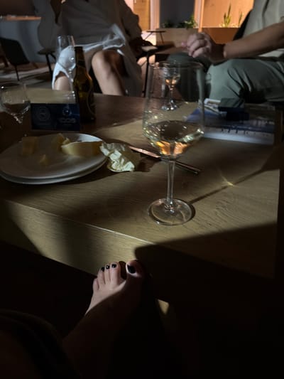 The image depicts a dimly lit, indoor scene, likely a lounge or restaurant, with elements of a casual social gathering. A wooden table occupies the foreground and mid-ground, reflecting ambient light and casting long shadows. On the table, two white plates are stacked, with one holding several pieces of cheese, suggesting a shared snack. A crumpled white napkin rests to the right of the plates. Adjacent to the plates is a tall wine glass containing a pale yellow liquid, likely white wine. To the left of the wine glass, a brown beer bottle stands upright. A blue and white rectangular object with "IQOS" branding is visible near the plates. In the background, out of focus, two individuals are present. One person, seated, wears a white robe, with their legs exposed. Another individual, seated to the right, wears green trousers and a beige top, with their hands clasped in front of them. The lower right portion of the image features a person's bare feet, with black nail polish on the toes, resting in the foreground. The environment appears to be dimly lit, possibly at dusk or night, with warm lighting highlighting certain objects on the table. The location within the city of Žunovnica, Bosnia and Herzegovina, cannot be determined from the visual information provided in the image.
