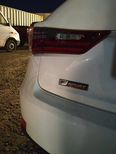 In Krtsanisi, Georgia, at night, a white Lexus F Sport sedan is parked on a gravel lot. The vehicle's rear is visible, with a prominent taillight and the "F SPORT" emblem. In the background, a white van and a dark-colored vehicle are parked near a corrugated metal structure. The ground is covered in gravel and appears wet, indicating recent rain. The lighting suggests artificial illumination from the environment.