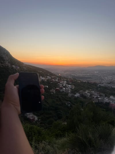 The image displays a panoramic view of a cityscape and surrounding natural landscape during sunset or dusk, likely in Tétouan, Morocco, viewed from an elevated position. The sky presents a striking gradient of colors, transitioning from a warm, intense orange and yellow along the horizon to a softer grey-blue higher up, indicating the late afternoon or early evening.

In the foreground, a person's hand is visible, holding a dark-colored smartphone. The phone is pointed towards the scenic view, suggesting the person is capturing a photo or video of the sunset over the city.

The midground features a dense urban area built on undulating hillsides and extending into a valley. Numerous buildings, predominantly white or light-colored, are scattered across the landscape, interspersed with abundant green vegetation and trees. Roads and structures wind through the city, with some faint lights already visible, indicating the onset of night. Distant mountains form a hazy silhouette against the colorful sky, defining the natural boundaries of the scene. On the immediate right, dark green foliage, including palm fronds, is prominent.

The overall scene conveys a serene and expansive view of the city as it settles under the twilight sky. No visible text is present in the image.