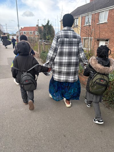 A group of five individuals, comprising two adults and three children, walk in a line along a grey asphalt pavement on a residential street in Rotherham, United Kingdom, under an overcast sky.
In the foreground, an adult, seen from behind, wears a black turban, a black and white plaid long-sleeved jacket, a long blue patterned skirt, and off-white clogs. This adult holds hands with two children, one on each side. The child to the adult's left wears a black hooded puffer jacket, dark trousers, and brown shoes, carrying a grey "AOTIANBAG" backpack. The child to the adult's right wears a black fur-lined hooded puffer jacket, grey trousers, and black and white Nike sneakers, carrying an olive green "AOTIANBAG" backpack.
Further ahead on the pavement, a second child, wearing a dark blue hooded jacket and dark trousers, walks in front of the primary group. Behind this child and further to the left, a second adult wears a long black coat with a fur-lined hood and dark trousers.
The environment consists of red brick terraced or semi-detached houses with white window frames and dark roofs, visible along the right side of the street. Bare deciduous trees and sparse vegetation line the property boundaries. A grey street light pole stands on the left side of the pavement.