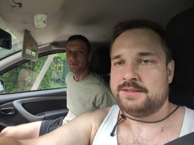 Two men are in the front seats of a car, possibly in Малая Листовка, Russia. The man in the foreground is sweating heavily and looking at the camera. He is wearing a white tank top and has a beard. The man in the background is looking out the window and appears to be wearing a light-colored t-shirt. The interior of the car is visible, including the dashboard, rearview mirror, and sun visor. The exterior of the car shows greenery and trees, suggesting a rural or suburban setting.