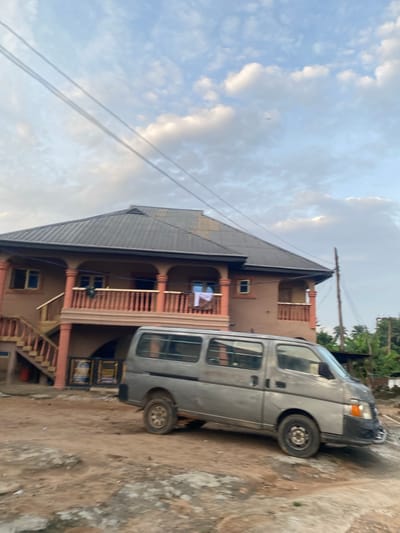 The image captures a static street scene in Enwan, Nigeria, under a partly cloudy, light blue sky traversed by two parallel power lines. A small, indistinct object, possibly an airplane, is visible in the upper right sky. The primary subject is a two-story building with a dark, possibly corrugated metal, gabled roof and reddish-brown walls, featuring multiple windows and two second-story balconies with reddish-orange railings and pillars. An exterior staircase, matching the balcony railings, ascends to the second floor on the left. A piece of white laundry hangs over the railing of the left balcony. In the foreground, a worn, light grey van, possibly a panel van, is situated on an unpaved, reddish-brown dirt road, partially obscuring the lower section of the building. The van's wheels are visibly dirty, and its front bumper appears damaged or discolored. To the right of the building, a rustic wooden utility pole stands amidst green tropical foliage, including palm trees. No individuals are visible in the scene.