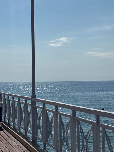This image depicts a scene on a pier overlooking a body of water, likely a lake, under a clear blue sky with scattered clouds. The pier has a wooden plank walkway and a white metal railing with a decorative pattern. A tall, grey pole, possibly a lamppost, stands vertically on the pier. The water is calm and reflects the sunlight, creating a shimmering effect. In the far distance, the horizon line where the water meets the sky is visible. The location is identified as the city of Koshkol', Kyrgyzstan. No people are visible in the image. There is a small dark bird perched on the railing in the mid-right portion of the image.
