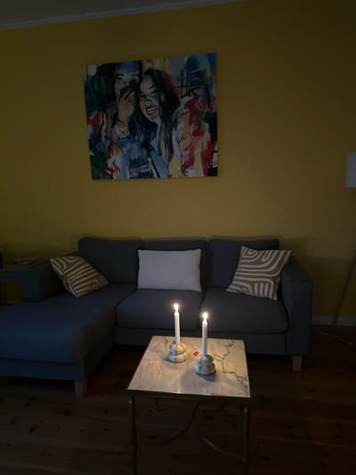 In Ammeldingen bei Neuerburg, Germany, a cozy living room is dimly lit by two lit candles on a marble-topped coffee table. The coffee table has a gold-toned metal frame. Behind the coffee table, a grey sectional sofa is arranged with two throw pillows: one white and one with a yellow and white abstract pattern. A large, colorful abstract painting depicting two laughing women hangs on a yellow wall above the sofa. The floor is made of warm-toned wooden planks.