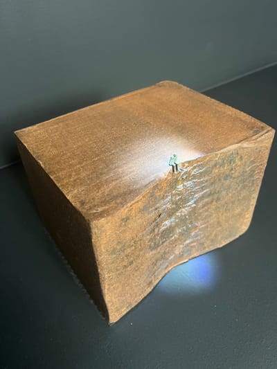 The image displays a miniature sculpture or diorama situated on a flat, dark grey surface against a dark grey wall background. The central object is a rectangular block, appearing to be composed of a coarse, oxidized material resembling rusted metal or weathered earth, characterized by a predominantly reddish-brown coloration with variations in texture and tone.
One corner of the block features a deeply carved, undulating depression, mimicking an eroded cliff face or natural cavern opening. At the upper edge of this depression, a single miniature human figure, rendered in teal-green attire, is positioned, appearing to look down into the void. A focused beam of white light emanates from the figure's vicinity, illuminating a section of the block's top surface and the upper part of the sculpted depression. Below the main block, within the shadowed area at the base of the depression, a faint, diffuse blue light is visible on the dark supporting surface.
The scene, an art piece or model, is located in the city of Galatina, Italy.