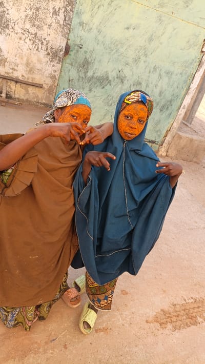 Two young individuals are captured in a vertical, eye-level shot, posing playfully outdoors during what appears to be daylight. Both have their faces, and possibly parts of their necks and hands, covered in a vibrant, granular orange substance, possibly a cosmetic mask or a traditional preparation like turmeric paste. Their eyes are clearly visible through the orange covering.

The person on the left wears a light blue head covering with a black and white patterned design. They are dressed in a loose-fitting, earthy brown garment, possibly an abaya or robe, which extends down to their ankles. A colorful, patterned fabric, possibly an underskirt or sarong with blues, greens, and browns, is visible beneath the brown garment. Their hands are clasped together in front of their chest, forming a shape that resembles a heart or a similar gesture, and one arm is draped around the shoulder of the person on the right.

The person on the right wears a dark blue head covering that appears to be part of a larger, flowing dark blue garment, likely a khimar or hijab, which covers most of their body. A distinct, colorful patterned fabric in oranges, blues, and browns is visible at the hem of their blue garment. They hold both hands up, forming a "peace" sign or V-shape with their fingers. Both individuals are looking towards the camera with a neutral to slightly smiling expression, though their expressions are somewhat obscured by the facial covering.

Their footwear consists of open-toed slide sandals. The person on the left wears light brown sandals, while the person on the right wears light green sandals.

The background features a textured wall on the left, which is off-white or light beige, exhibiting signs of wear, cracks, and discoloration. A wooden plank or piece of timber rests against this wall. On the right, there is a large, flat, light green or mint-colored surface, possibly a weathered metal door or gate, showing scratches and some rust. The ground is a light brown or beige, dusty surface, possibly dirt or concrete. The overall setting suggests a rustic or aged outdoor environment. The lighting is bright and even, indicating either an overcast sky or diffuse daylight.