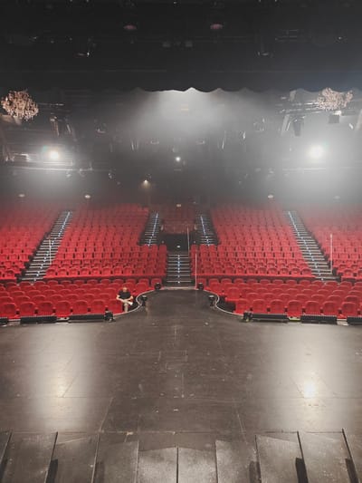 A wide, high-angle shot captures an empty theater with rows of plush red seats. The seats are arranged in tiers, with illuminated staircases providing access. In the center of the frame, on the stage floor, a lone man sits casually, looking towards the audience. The stage is dark and bare, with professional lighting fixtures visible overhead. The overall ambiance is one of anticipation, with the empty seats awaiting a performance. The location is the Room of Kirrwiller, France.