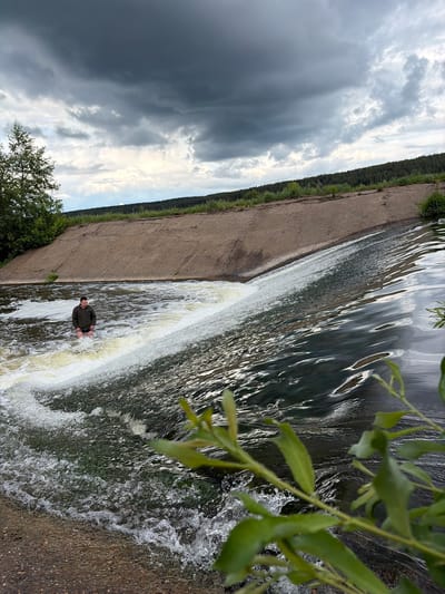 A man stands at the base of a small artificial waterfall in what appears to be Nizhnii Vishur, Russia. The waterfall is created by a dam or weir structure, and the man is standing in the cascading water. The background shows a concrete structure with vegetation along the edge, along with a treeline and a cloudy sky. A leafy branch is in the extreme foreground, partially obscuring the upper right.